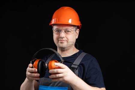 Téléchargez les photos : Bâtisseur dans un casque de protection avec des écouteurs anti-bruit dans ses mains sur un fond noir, espace de copie pour le texte. - en image libre de droit