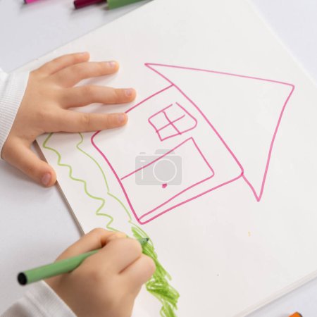 Foto de Pequeño niño dibujando con rotuladores una casa con hierba en un cuaderno de bocetos - Imagen libre de derechos