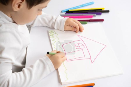 Foto de Pequeño niño dibujando con rotuladores una casa con hierba en un cuaderno de bocetos - Imagen libre de derechos