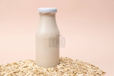 Oat milk in a glass bottle among dry oatmeal flakes on a beige background