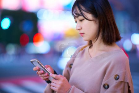 Téléchargez les photos : Jeune femme regardant smartphone dans le centre-ville la nuit - en image libre de droit