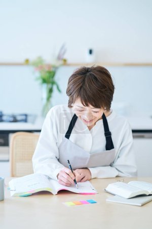Photo for A woman in an apron studying by text at kitchen - Royalty Free Image