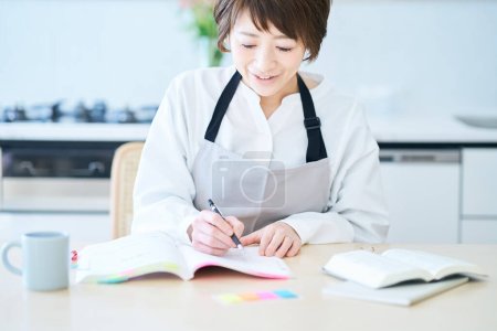Téléchargez les photos : Une femme dans un tablier étudiant par texte à la cuisine - en image libre de droit