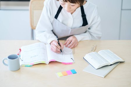 Téléchargez les photos : Une femme dans un tablier étudiant par texte à la cuisine - en image libre de droit