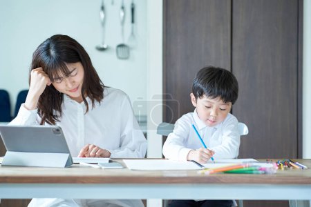 Téléchargez les photos : Un enfant jouant dans la chambre et une mère avec une expression fatiguée après avoir travaillé à côté - en image libre de droit