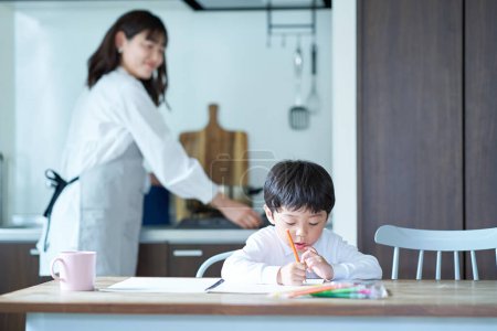 Foto de Un niño que dibuja y una madre que vigila mientras hace tareas domésticas en casa - Imagen libre de derechos