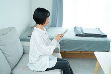 Téléchargez les photos : Femme assise sur un canapé et utilisant un smartphone dans la chambre - en image libre de droit