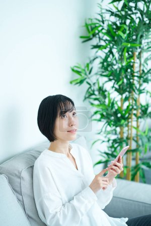 Téléchargez les photos : Femme assise sur un canapé et utilisant un smartphone dans la chambre - en image libre de droit