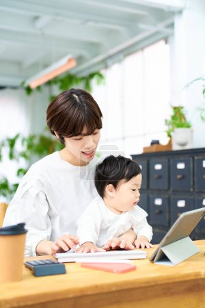Photo for A woman holding a baby and operating a computer indoors - Royalty Free Image