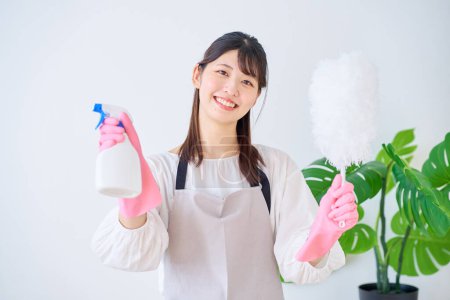 Photo for Young woman in an apron holding cleaning tools indoors - Royalty Free Image