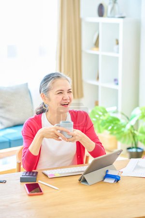Téléchargez les photos : Femme âgée souriante prenant une pause tout en travaillant à la maison avec une tasse - en image libre de droit