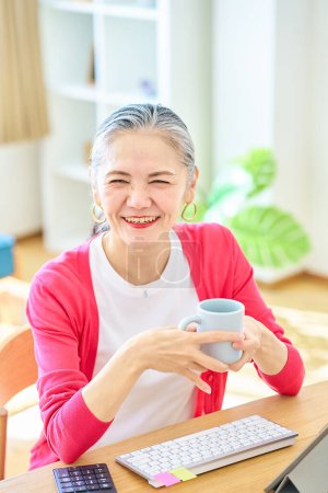 Téléchargez les photos : Femme âgée souriante prenant une pause tout en travaillant à la maison avec une tasse - en image libre de droit