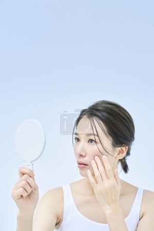 Téléchargez les photos : Une femme avec une expression anxieuse vérifiant son état de la peau avec un miroir de main - en image libre de droit