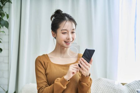Téléchargez les photos : Jeune femme souriante portant des vêtements de salon opérant un smartphone - en image libre de droit
