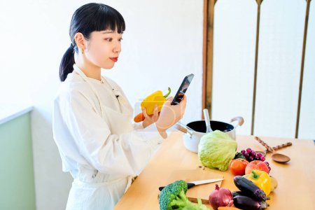 Téléchargez les photos : Jeune femme ramasser des légumes et smartphone dans la cuisine - en image libre de droit