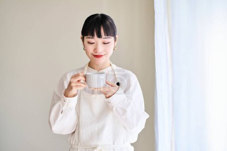 Photo for A young woman in an apron taking a break by the window in the room - Royalty Free Image