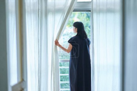 Mujer joven abriendo las cortinas de encaje y mirando por la ventana