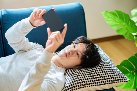Photo for Young man lying on a sofa bed holding a smartphone in the room - Royalty Free Image