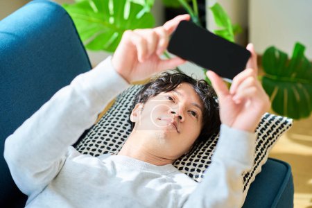 Young man lying on a sofa bed holding a smartphone in the room