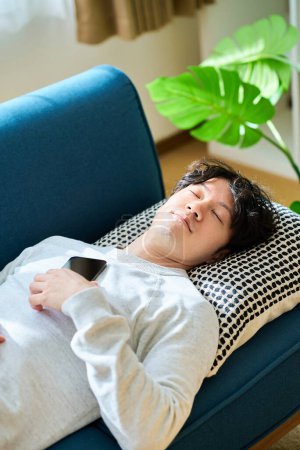 Téléchargez les photos : Jeune homme couché sur un canapé-lit tenant un smartphone dans la chambre - en image libre de droit