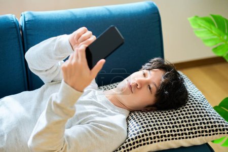 Junger Mann liegt auf Schlafcouch und hält Smartphone im Zimmer