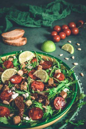 Ensalada de verduras frescas: rúcula, tomates cherry frescos y secos, piñones y queso sobre fondo verde, vertical