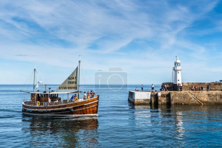 Téléchargez les photos : Mevagissey, Cornouailles, Royaume-Uni - 16 août 2023 : Le phare et l'entrée du port de Mevagissey sur la côte sud de Cornouailles. Un bateau touristique revient d'un tour. - en image libre de droit