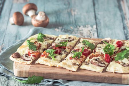 Photo for Freshly baked flammkuchen, traditional french tarte flambee or german pizza in a vegetarian recipe with mushrooms, cream cheese, tomatoes and arugula, on light green wooden board and table. - Royalty Free Image