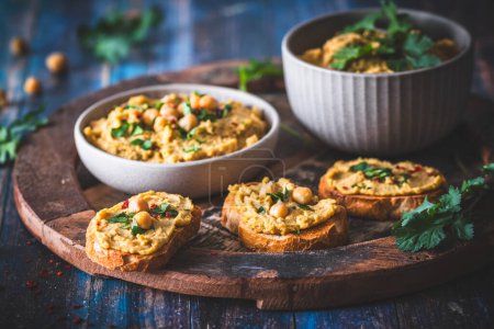 Hummus, chickpea puree in a bowl and on toasted bread slices on blue wooden background