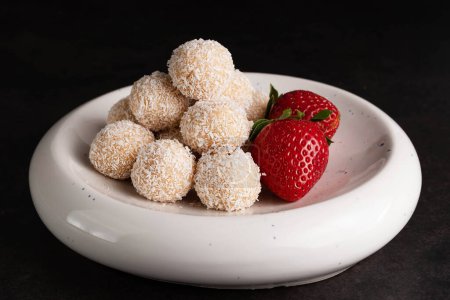 Téléchargez les photos : Sur fond sombre, bonbons à la noix de coco et fraises dans une assiette légère. - en image libre de droit