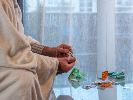 Foto de Un hombre enfermo con el virus envuelto en una manta con un termómetro en la mano, tabletas y toallitas nasales para una secreción nasal en la mesa - Imagen libre de derechos