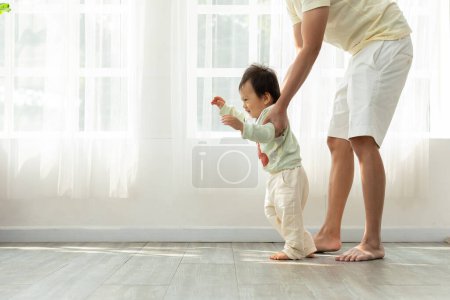 Foto de Adorable bebé asiático niño aprendiendo a caminar con papá ayudando en casa. Pequeño niño mirando al frente a caminar paso a paso y padre apoyando a aprender y desarrollar habilidades al lado. Primeros pasos - Imagen libre de derechos
