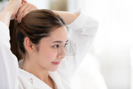 Foto de Hermosa mujer joven asiática atar el cabello de nuevo antes de la ducha o aplicar crema o loción en la cara. Belleza femenina agarrar sus pelos después de despertar con felicidad y emocional positiva. Concepto de autocuidado - Imagen libre de derechos
