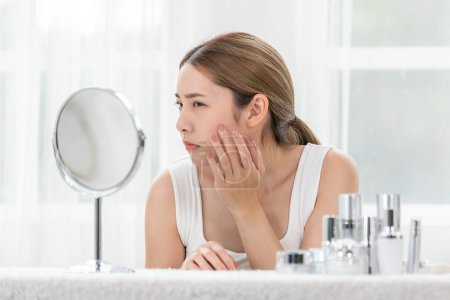 Asian young woman looking in mirror worry about pores facial skin problem with acne and dry skin. Self care and Skin care concept