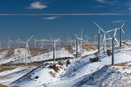 Foto de Turbinas eólicas en una granja de molinos de viento para la producción de energía alternativa que se muestra en las montañas Tehachapi cubiertas de nieve en el sur de California. Las montañas de San Gabriel se ven a lo lejos. - Imagen libre de derechos