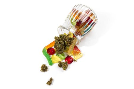 Foto de Varias gomitas y cogollos de marihuana medicinal se cayeron del frasco de vidrio en relieve. Sobre un fondo blanco. Un montón de espacio vacío, vista superior - Imagen libre de derechos