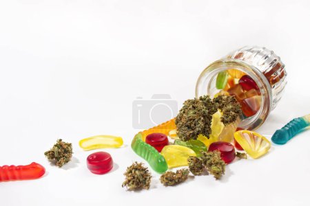 Varias gomitas y cogollos de marihuana medicinal se cayeron del frasco de vidrio en relieve. Sobre un fondo blanco. Un montón de espacio vacío