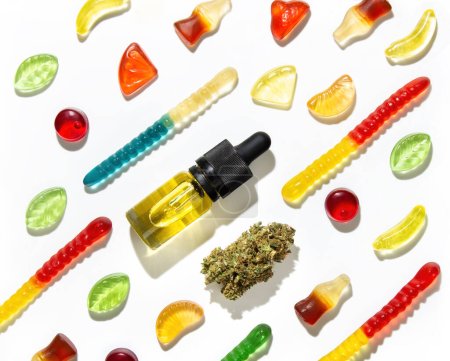 Among Chewing marmalade of different shapes, tastes and colors, a dried cannabis bud and bottles of marijuana seed oil lie in a pattern on a white background in the center.