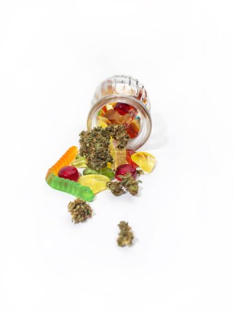 Foto de Varias gomitas y cogollos de marihuana medicinal se cayeron del frasco de vidrio en relieve. Sobre un fondo blanco. Un montón de espacio vacío - Imagen libre de derechos