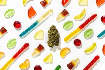 Foto de Entre gomitas de diferentes formas, sabores y colores, un brote de cannabis seco se encuentra sobre un fondo blanco en el centro. - Imagen libre de derechos