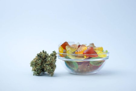 Foto de Surtido de gomitas en un tazón de vidrio pequeño junto a varios brotes de marihuana medicinal. Sobre un fondo azul pálido, un montón de espacio vacío - Imagen libre de derechos
