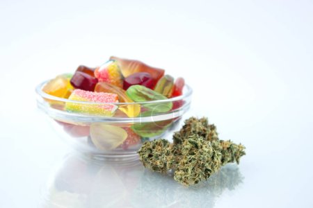 Assorted different gummy candies in a small glass bowl, near in the foreground several buds of dried medical marijuana, reflected on the glass