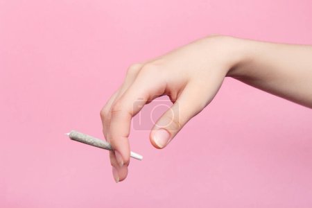 Foto de La mano de una mujer sostiene un porro con marihuana medicinal con los dedos. Sobre un fondo rosa - Imagen libre de derechos