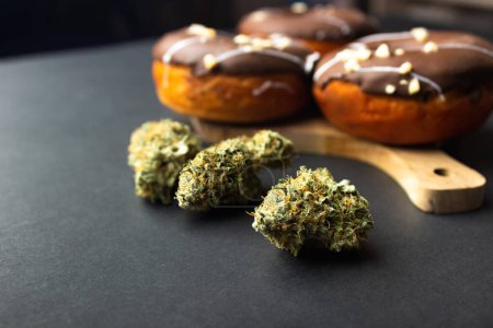 Trockene Knospen medizinischen Marihuanas aus nächster Nähe, im Hintergrund Donuts, die mit Schokoladenglasur überzogen und mit Nusskrümeln bestreut sind. Auf schwarzem Hintergrund