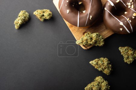 Schokoladenüberzogene Donuts mit Haselnussbelag liegen auf einem Holzbrett zwischen trockenen Knospen medizinischen Marihuanas. Auf schwarzem Hintergrund, flach