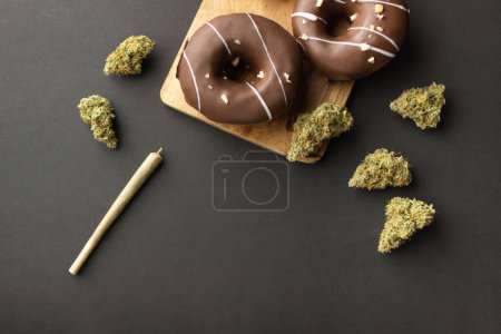 Schokoladenüberzogene Donuts mit Haselnussbelag liegen auf einem Holzbrett zwischen trockenen Knospen medizinischen Marihuanas neben einem Joint. Auf schwarzem Hintergrund, flach