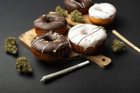 Donuts mit Schokolade und weißem Zuckerguss liegen auf Holzbrettern zwischen trockenen Knospen medizinischen Marihuanas, neben zwei Joints. Auf schwarzem Hintergrund