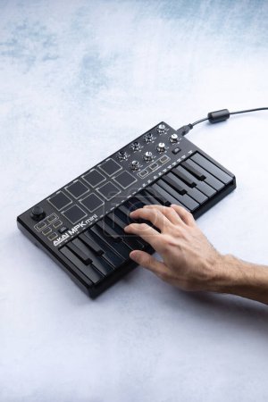 Foto de La mano de un hombre toca las teclas de un teclado midi negro AKAI mpk mini mk2, sobre un fondo blanco y azul. Vista lateral - Imagen libre de derechos