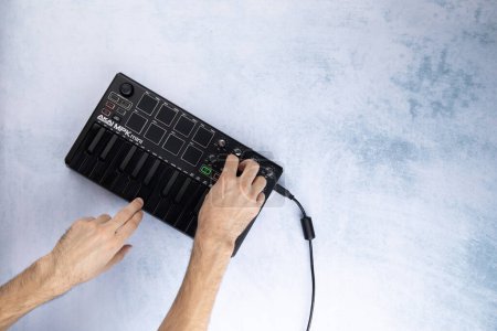 Foto de Las manos masculinas tocan las teclas y giran el codificador del teclado midi Akai mpk mini mk2 negro, sobre un fondo blanco y azul. Vista superior - Imagen libre de derechos