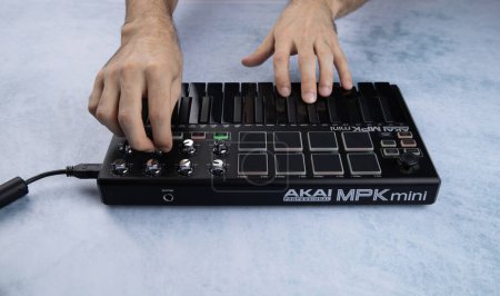 Téléchargez les photos : Une main d'homme joue les touches d'un mini clavier midi noir Akai mpk mk2, sur fond blanc et bleu - en image libre de droit